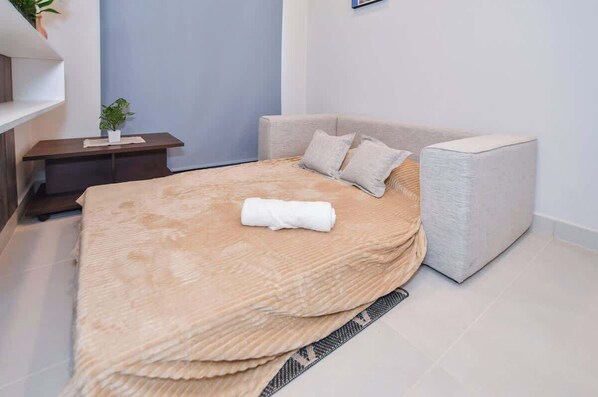 Una cama bien hecha con una manta color beige, una mesita de noche con una planta y un otoman gris.