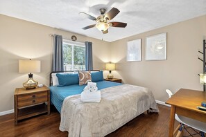 Une chambre à coucher comprenant un lit, un ventilateur de plafond, des lampes de chevet, une fenêtre avec des rideaux et des œuvres d’art encadrées accrochées au mur.