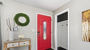 Un couloir avec une porte rouge, une console sur laquelle sont posés un vase et des fleurs, une guirlande accrochée au mur et une porte dotée d’un panneau de verre.