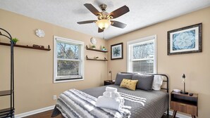 Une chambre à coucher comprenant un lit, un ventilateur de plafond, une table de chevet, une fenêtre et une étagère avec des objets décoratifs.