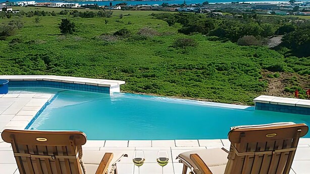 Une piscine bordée de deux chaises en bois et d’une table, le tout entouré d’une végétation luxuriante et offrant une vue sur la mer.