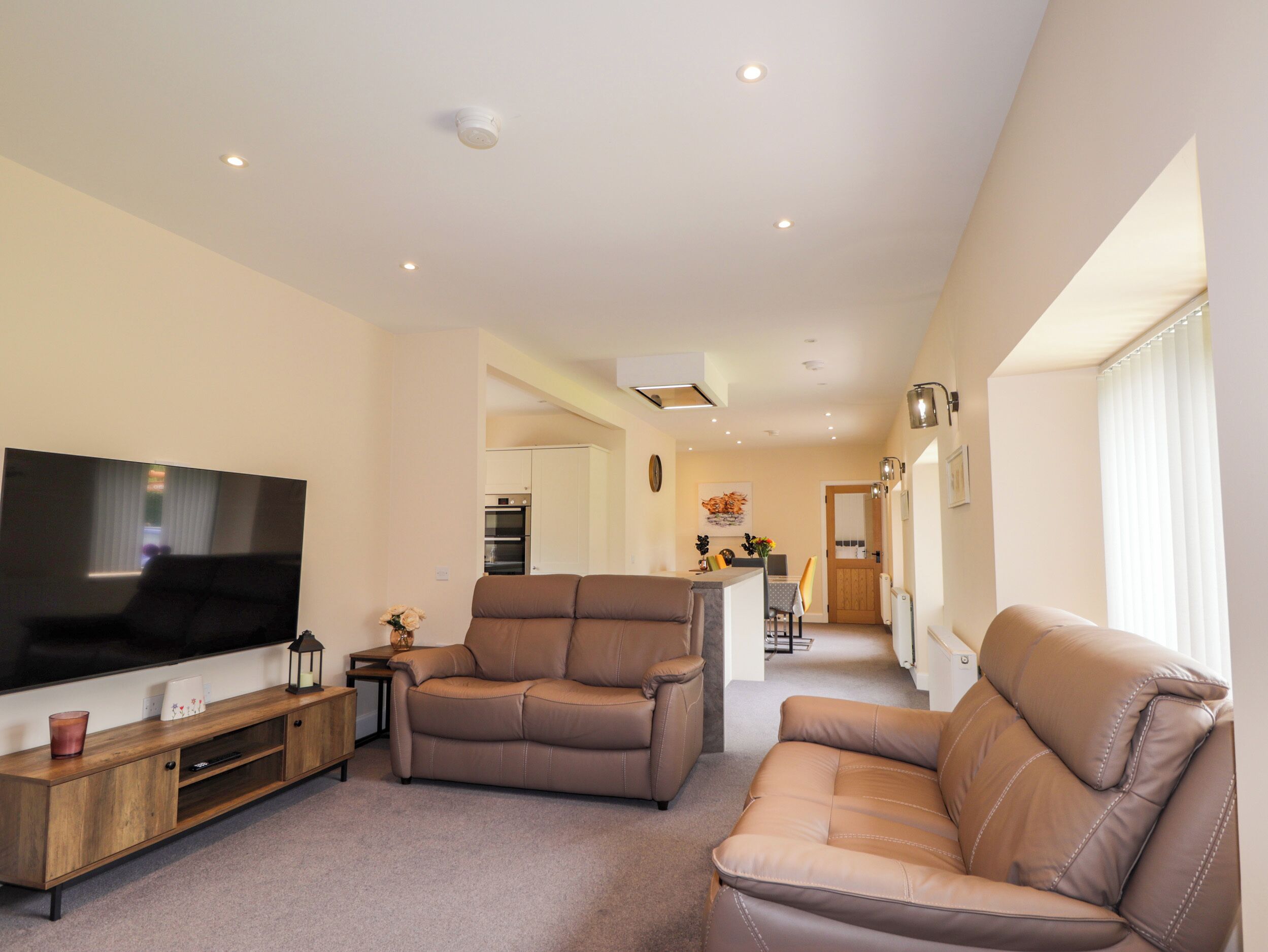 A living room with a flat-screen TV, two leather sofas, a wooden entertainment unit, and recessed ceiling lighting.