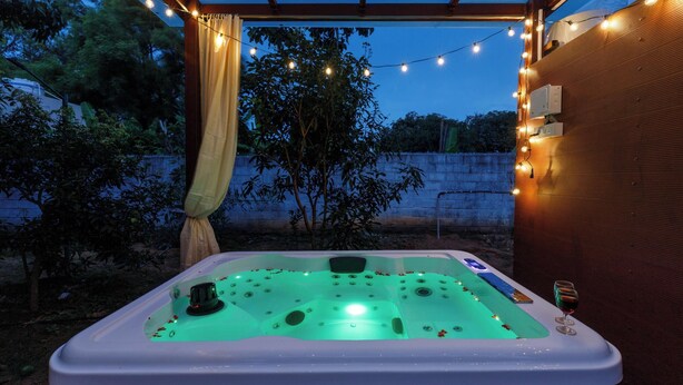 A hot tub under a pergola with string lights, surrounded by trees and a wooden wall.