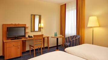 Een hotelkamer met een bed, een bureau met een televisie, een stoel, een klein tafeltje met een lamp en een raam met gordijnen.