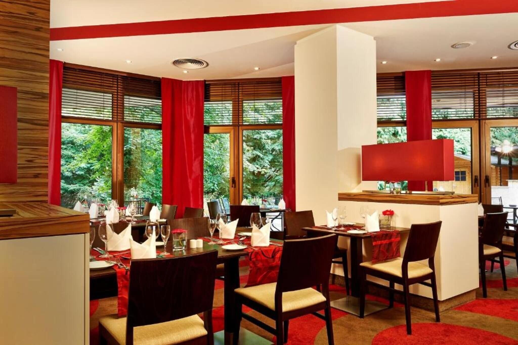 Un coin repas avec une table dressée pour quatre personnes, des rideaux rouges et de grandes fenêtres donnant sur un paysage verdoyant.