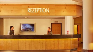 Ein Empfangsschalter mit dem Schild 'RECEPTION' darüber, zwei Angestellte an der Rezeption, ein Fernsehbildschirm und ein Holzboden im Empfangsbereich.