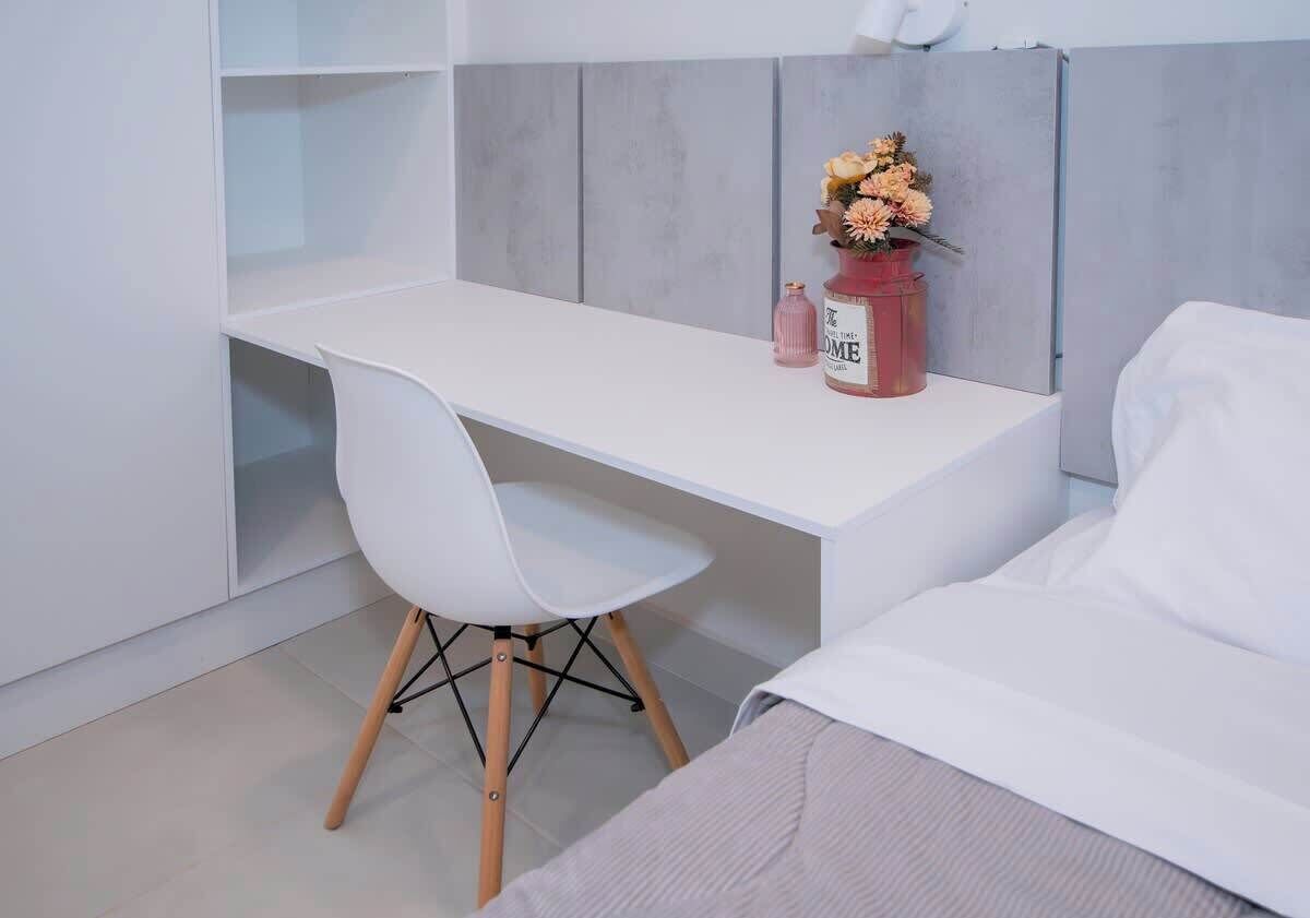 Une chambre moderne avec un lit blanc, un bureau blanc et une chaise. On peut voir un petit vase rose avec des fleurs sur le bureau.