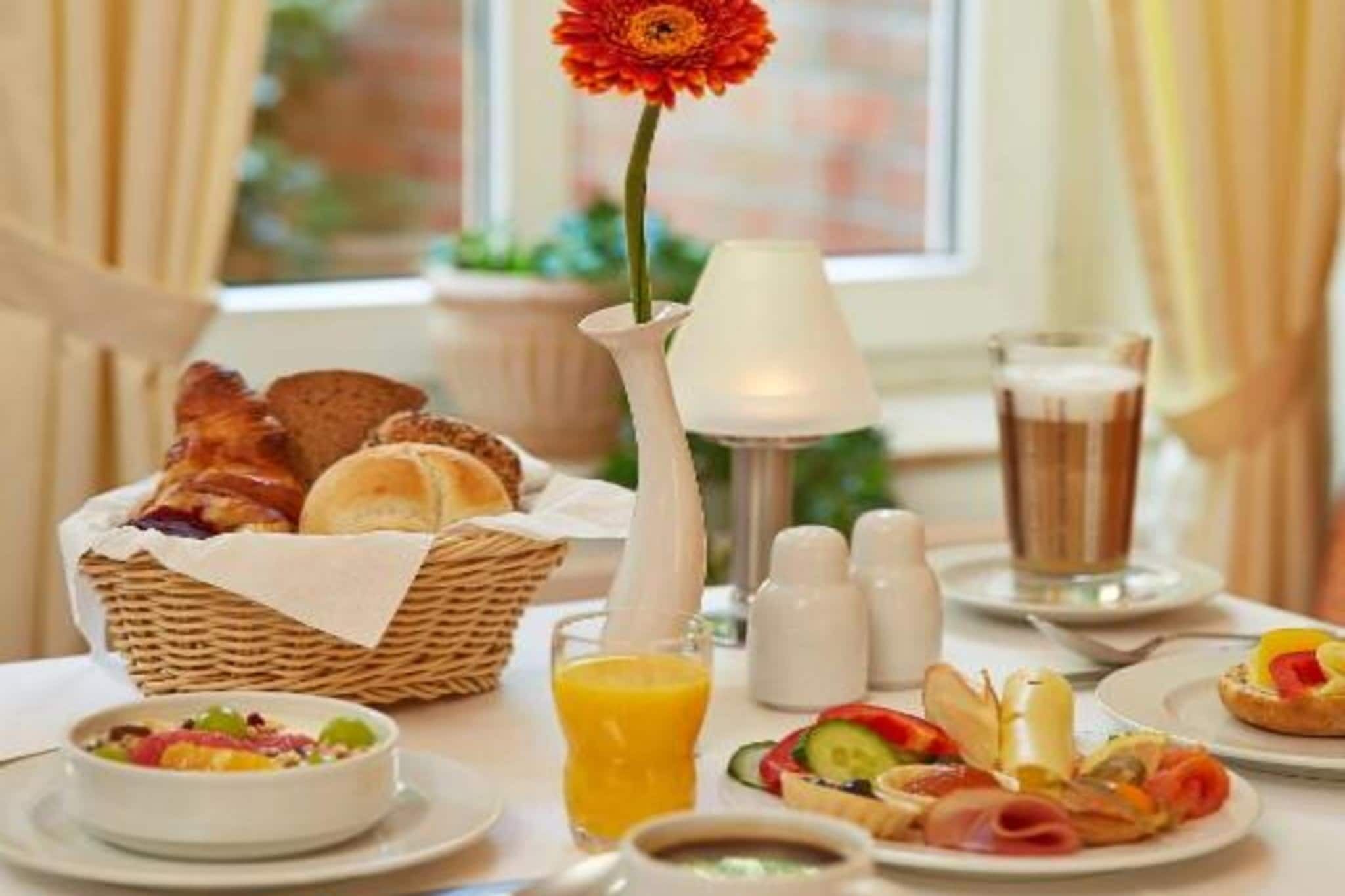 A breakfast setting with a basket of bread, a flower vase, a glass of orange juice, and plates with various foods.