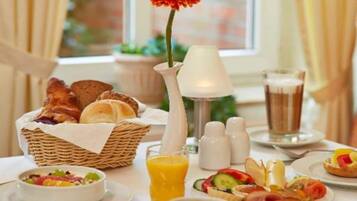 A breakfast setting with a basket of bread, a flower vase, a glass of orange juice, and plates with various foods.