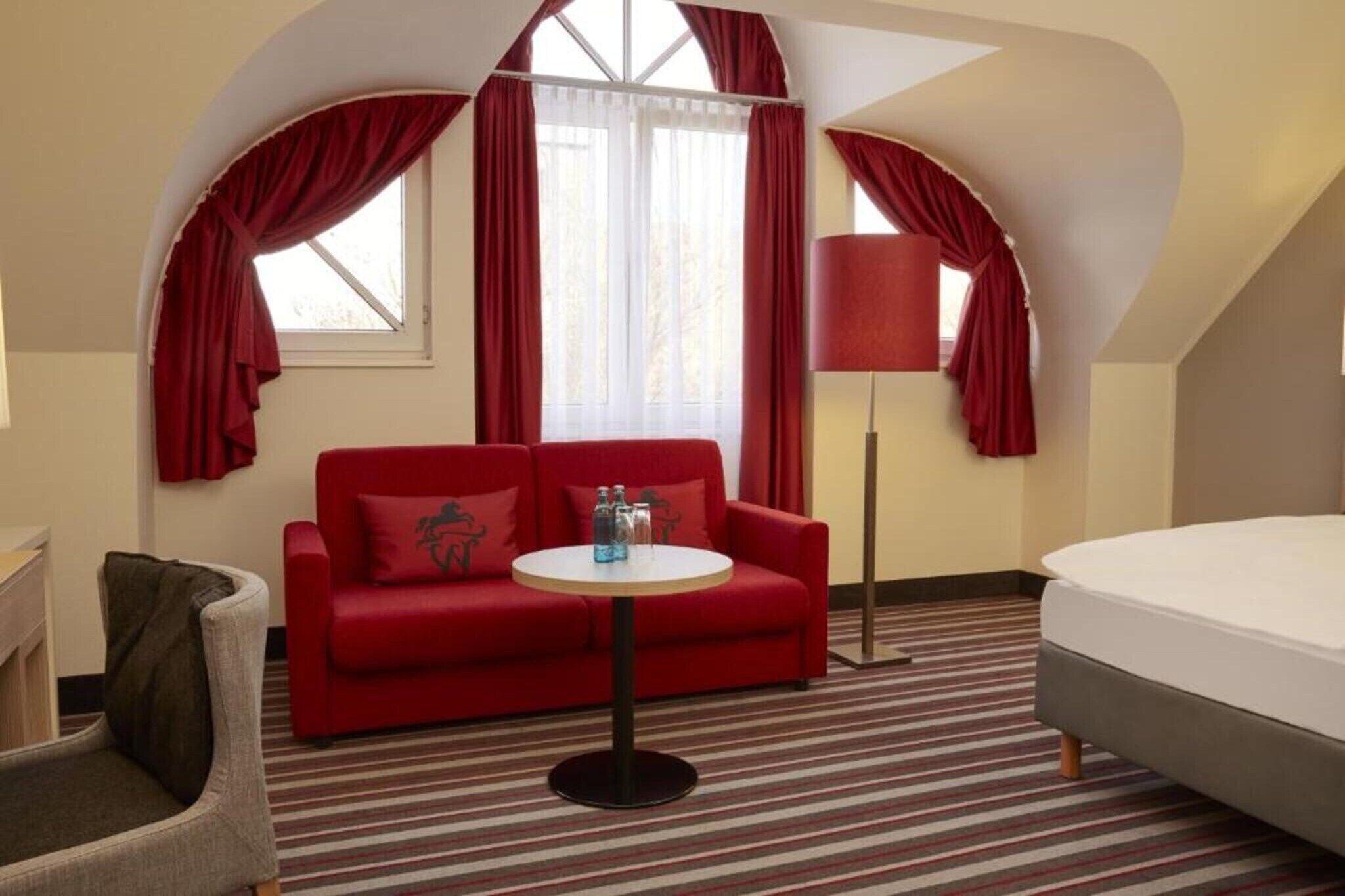 A hotel room with a red sofa, a small round table with bottles, a lamp, a bed, and a chair.