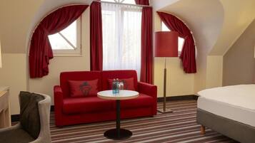A hotel room with a red sofa, a small round table with bottles, a lamp, a bed, and a chair.