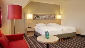 A hotel room with a bed, a nightstand, a red sofa, a round table with two bottles, and a wall art piece. - Sunday Hotel Stade Widukind Superior Room (Stade)