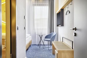 Ein Hotelzimmer mit Bett, kleinem Tisch, Stuhl, an der Wand montiertem Fernseher und einem Fenster mit Vorhängen.