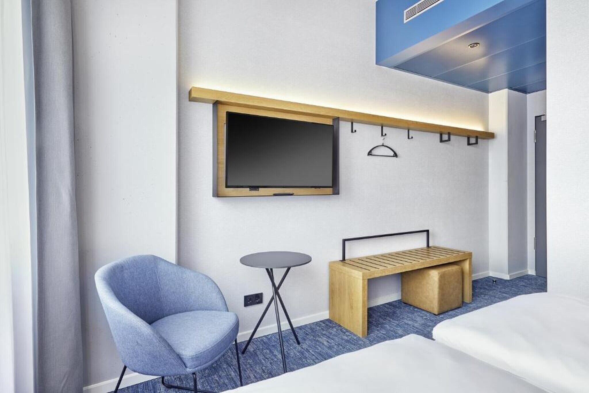 Une chambre d’hôtel moderne avec un lit, un fauteuil bleu, une petite table ronde, un banc en bois, un téléviseur fixé au mur et un luminaire au plafond.