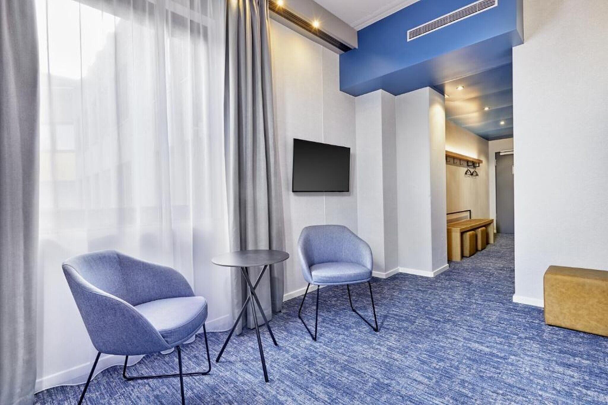 Une chambre d’hôtel avec deux chaises, une petite table, un téléviseur à écran plat et un couloir recouvert d’une moquette bleue.