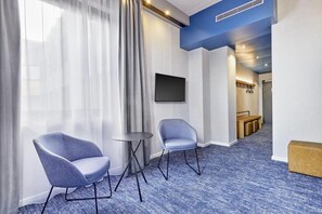 Een hotelkamer met twee stoelen, een tafeltje, een flatscreen televisie en een blauw tapijt in de gang.