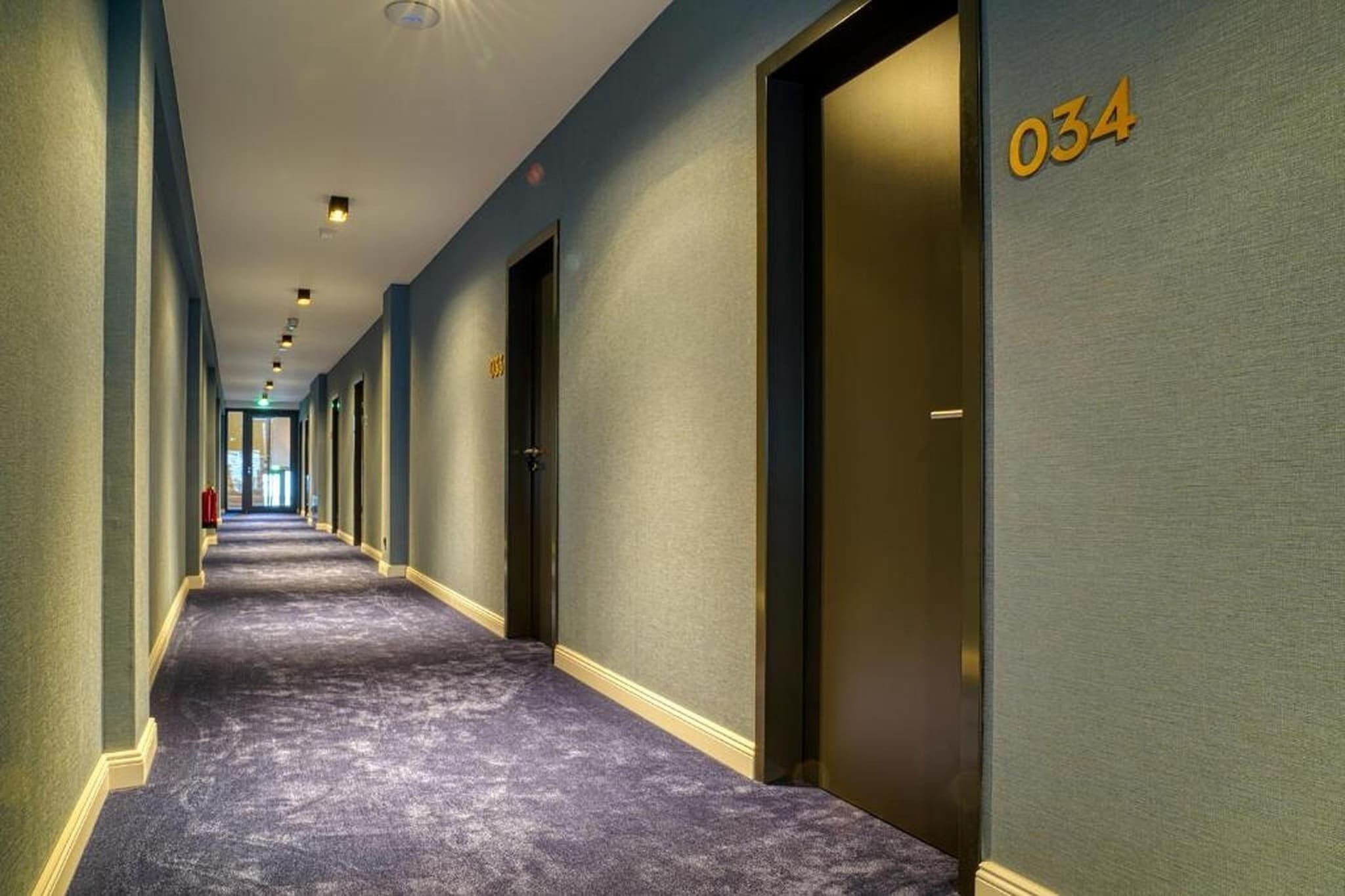 A hotel hallway with a carpeted floor, doors on the right, and a number '034' on the wall.