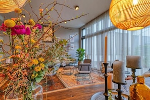 A room with a wooden floor, a large window, a table with a vase of flowers, and a candle holder.