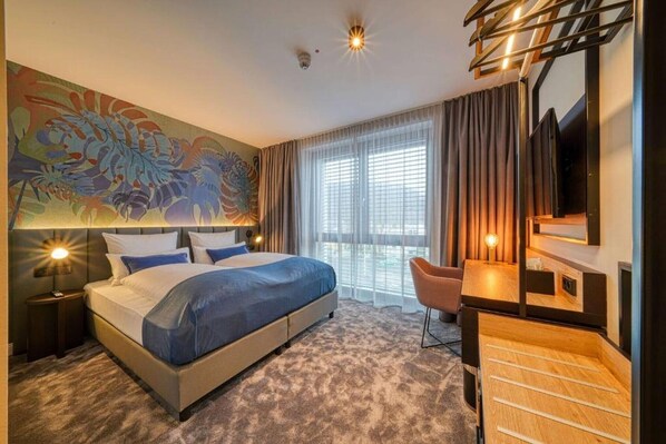 A hotel room with a large bed, a desk, a chair, a TV, and a balcony with curtains.