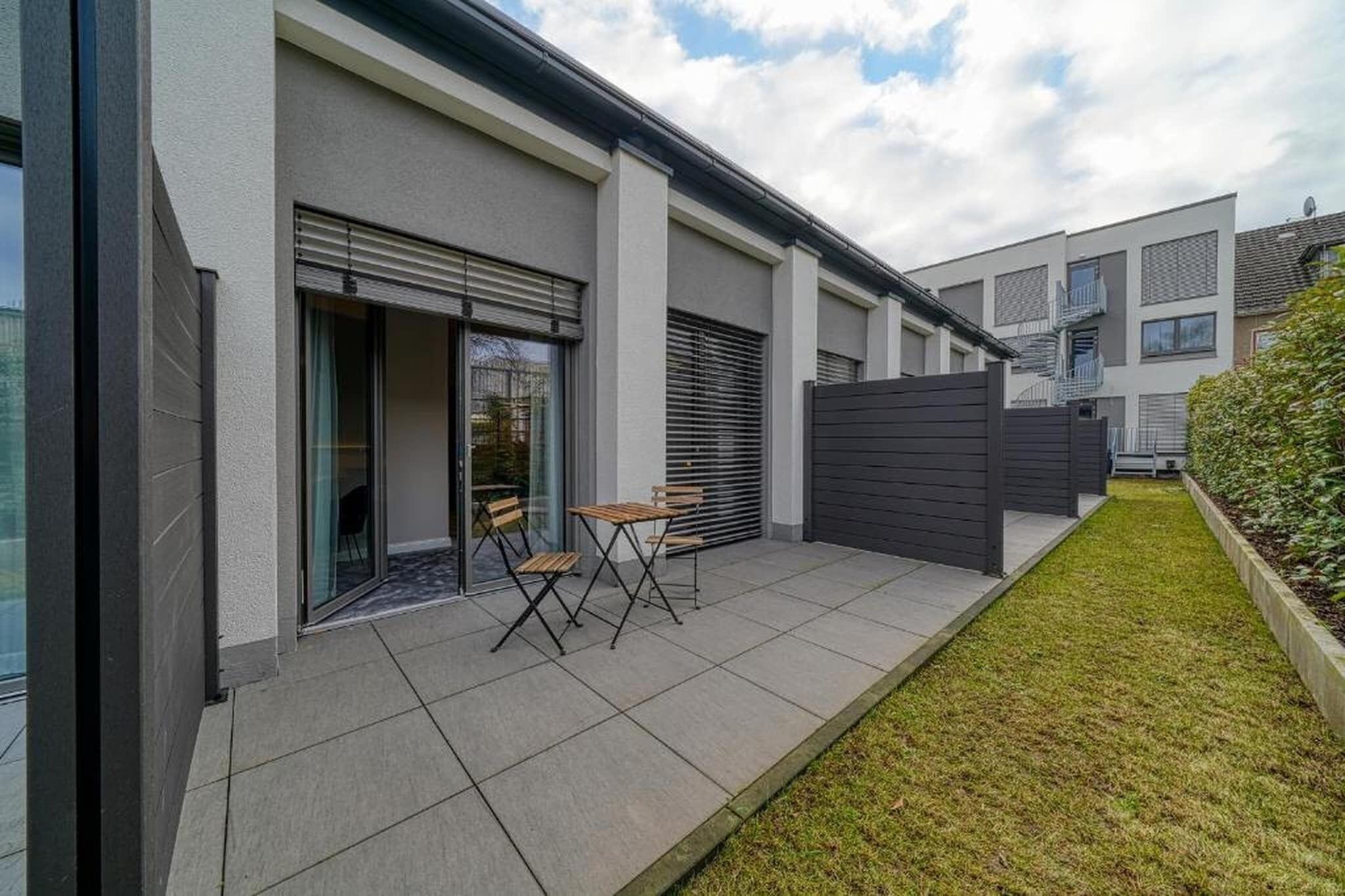 Ein modernes Wohngebäude mit einer Terrassenfläche, die einen Tisch und Stühle, eine Schiebetür und einen umzäunten Garten zeigt.