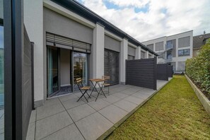 A modern residential building with a patio area featuring a table and chairs, a sliding glass door, and a fenced garden.