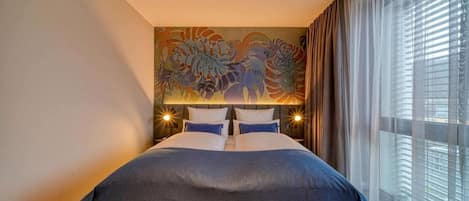 A hotel room with a large bed, a painting on the wall, and a window with curtains.