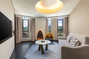 Habitación de hotel moderna con chimenea, sofá, mesa de centro y ventanas amplias con vistas al campo.
