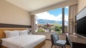 Une chambre d’hôtel comprenant un lit, un bureau avec une télévision, une chaise et une grande fenêtre donnant sur un temple et des montagnes.