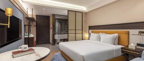 A modern hotel room with a large bed, bedside tables, a round table, a television, and a bathroom visible through a glass door.