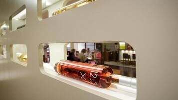 Un intérieur moderne où un objet cylindrique est exposé dans une vitrine transparente ; il pourrait s’agir d’une montre ou d’un objet décoratif. On aperçoit, en arrière-plan, le reflet d’un espace public.