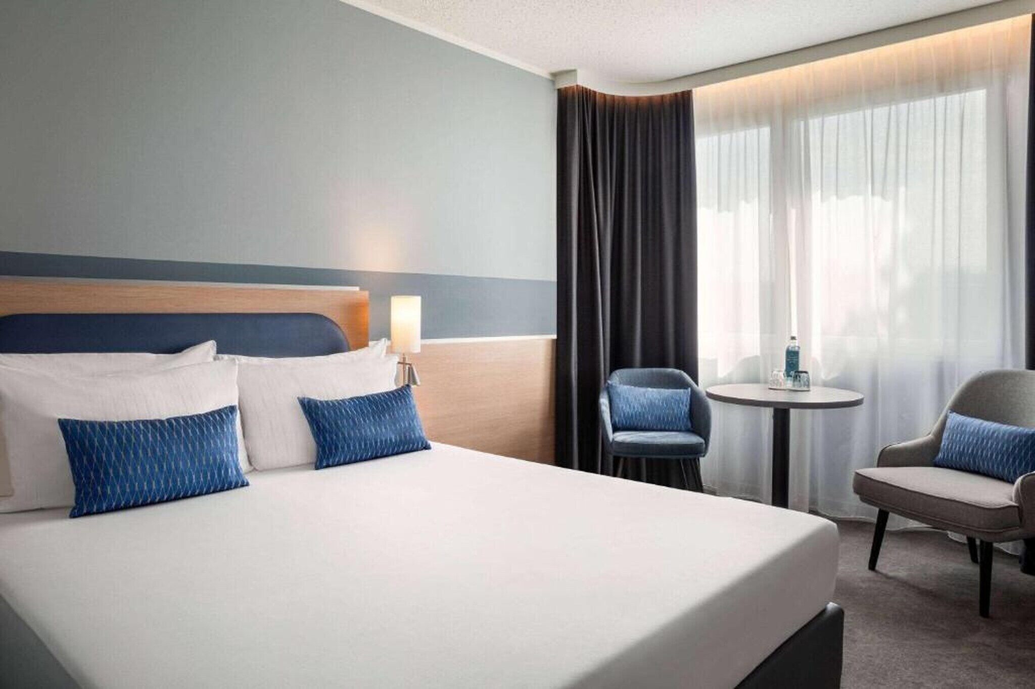 Habitación de hotel con una cama grande, dos sillas, una mesita y una ventana con cortinas.