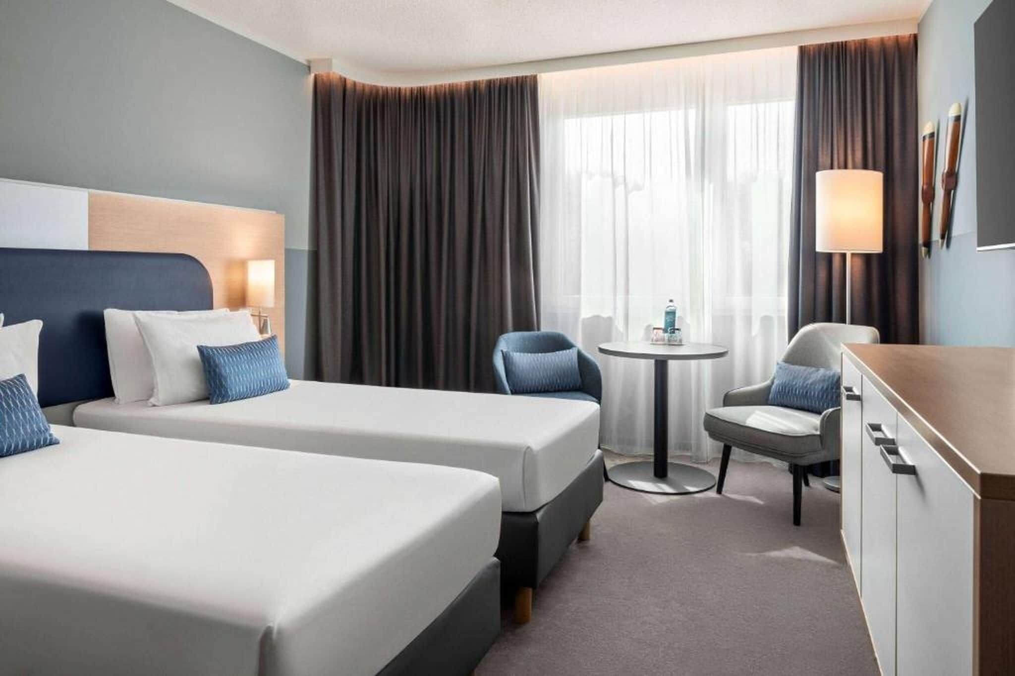 Habitación de hotel con dos camas, una mesita, una silla y un armario.