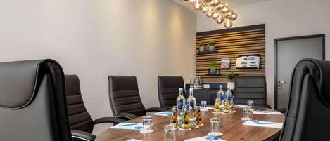 Una sala de conferencias con una mesa redonda, sillas de cuero negro, una lámpara de araña moderna y una pared de madera decorativa con estanterías.