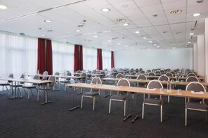 Un salón de conferencias con filas de mesas y sillas, ventanas amplias con cortinas e iluminación empotrada en el techo.
