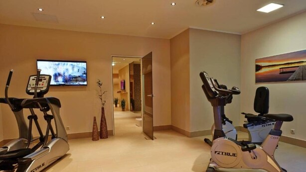 Un gimnasio en casa con dos bicicletas estáticas y una cinta de correr, un televisor en la pared y un pasillo que conduce a otra habitación.