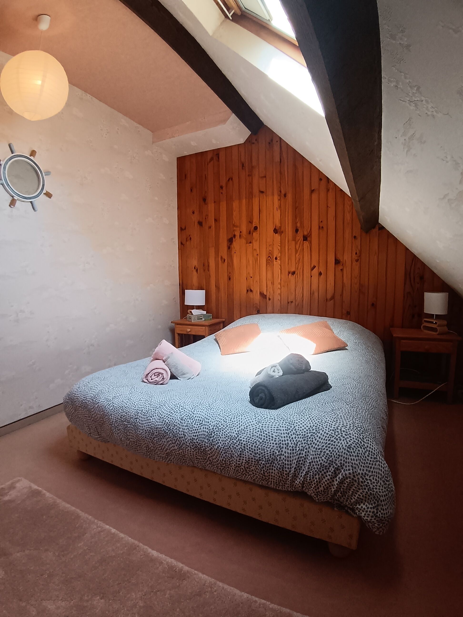 A bedroom with a sloped ceiling, wooden panel walls, a bed with patterned bedding, two bedside lamps, and a round mirror on the wall.