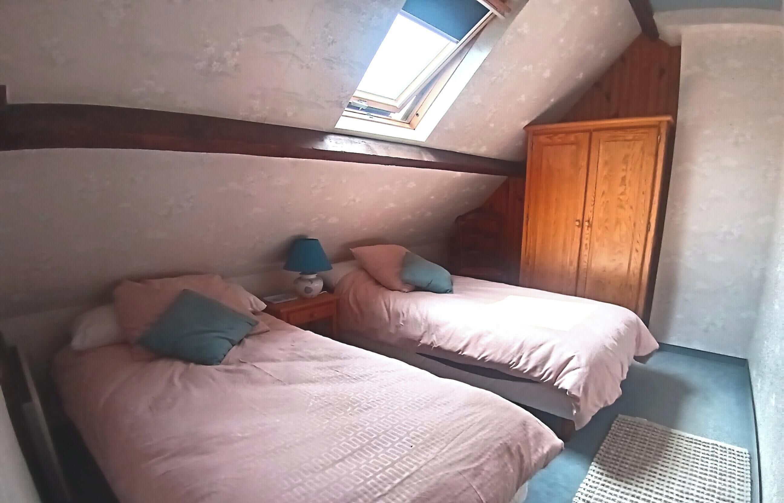A room with two beds, a wooden wardrobe, a small table with a lamp, and a skylight.