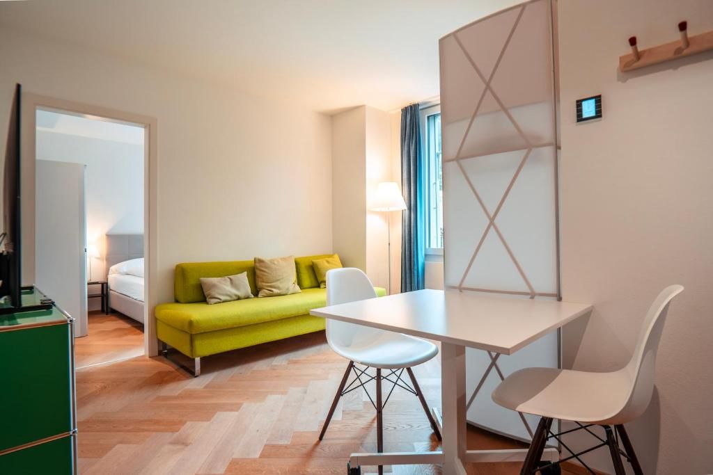 Een compacte kamer met een bed, een groene bank, een witte tafel met twee stoelen en een aan de muur bevestigde televisie.