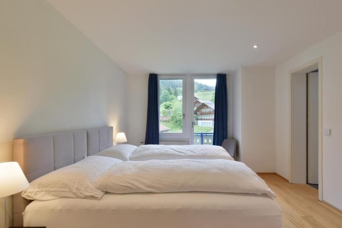 Une chambre avec un grand lit, une fenêtre avec des rideaux, une table de chevet avec une lampe et une porte sur le côté droit.