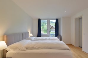 A bedroom with a large bed, a window with curtains, a nightstand with a lamp, and a door to the right.