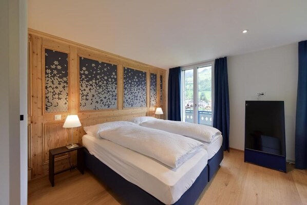 A double bed with a wooden headboard, a bedside table with a lamp, and a large window with curtains.