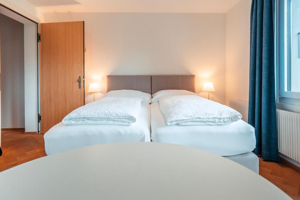 Ein Doppelzimmer mit zwei Betten, einer Holztür, einem fenster mit Vorhängen und einem runden Tisch im Vordergrund.