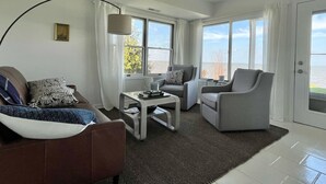 Una sala de estar moderna con un sofá, un sillón y una mesa de centro, ventanas amplias con vista al mar y una puerta que da al exterior.