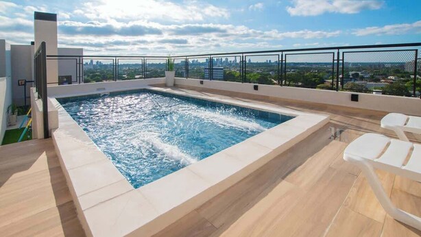 En pool på taket med utsikt över staden, omgiven av ett metallstaket, och med ett område med sittplatser.