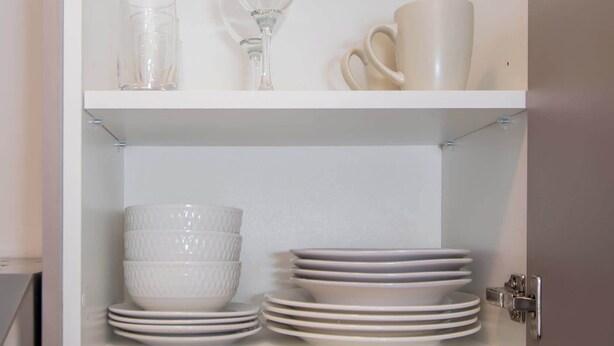 Un mueble blanco con estantes que contienen vasos de vidrio, una copa de vino y tazas blancas.