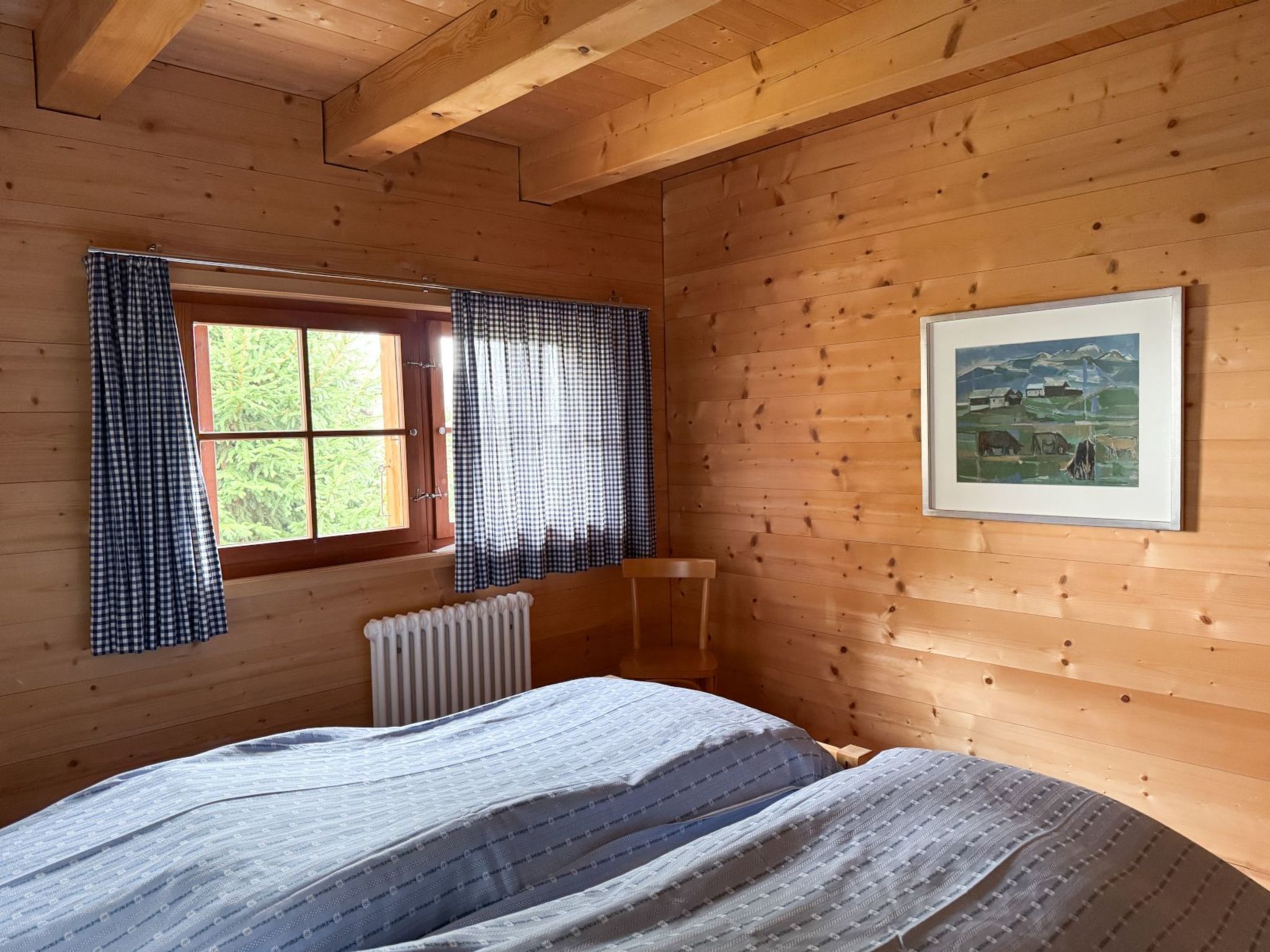 Ein Zimmer in einer Holzhütte mit einem Bett, einem Vorhangfenster, einem Heizkörper und einem gerahmten Bild an der Wand.