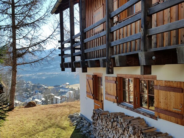A chalet with a wooden balcony, a stone wall, and a view of a valley.