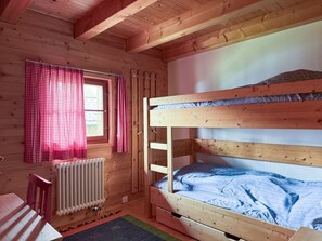 A wooden cabin room with bunk beds, a window with red curtains, a radiator, and a small chair.