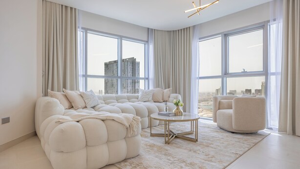 A modern living room with a sectional sofa, a round coffee table, and a chair, large windows with curtains, and a city view. - Bloomfields 2BR plus Maid in Radiant  (Abu Dhabi)