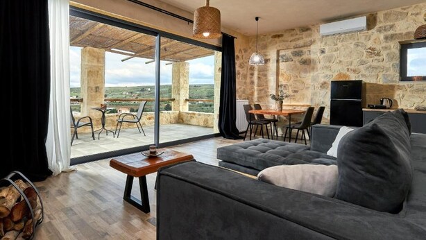 A modern living room with a sofa, dining table, and chairs, a stone wall, and a large glass door leading to an outdoor patio. - ALAGNI CRETAN RESORT (Alagni)